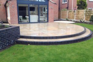  Natural stone patio in a circular design with brick built raised bed and steps to a lawn.