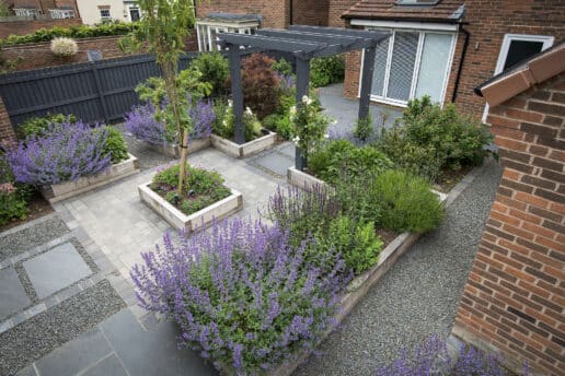 Modern garden design featuring purple flowering plants, raised beds, grey patio paving and contemporary pergola structure.
