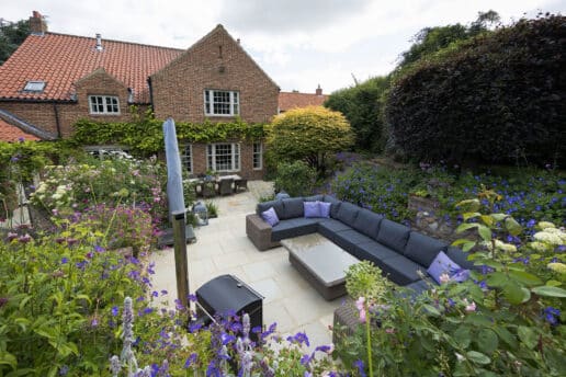 Completed terraced garden design with patio seating area, planted borders with purple flowers, and brick heritage cottage.