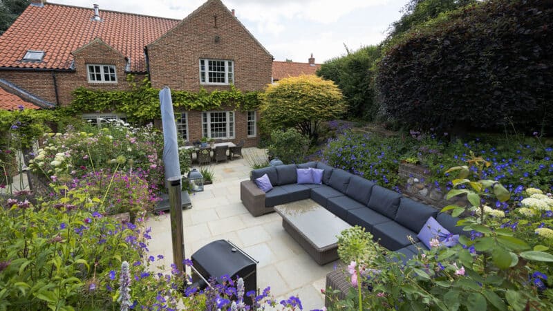 Completed terraced garden design with patio seating area, planted borders with purple flowers, and brick heritage cottage.